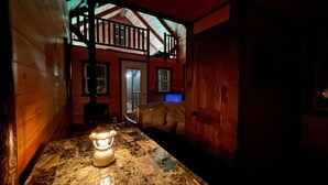 Un interior de madera tenuemente iluminado, con una mesa, un banco y una puerta que da a otra habitación.
