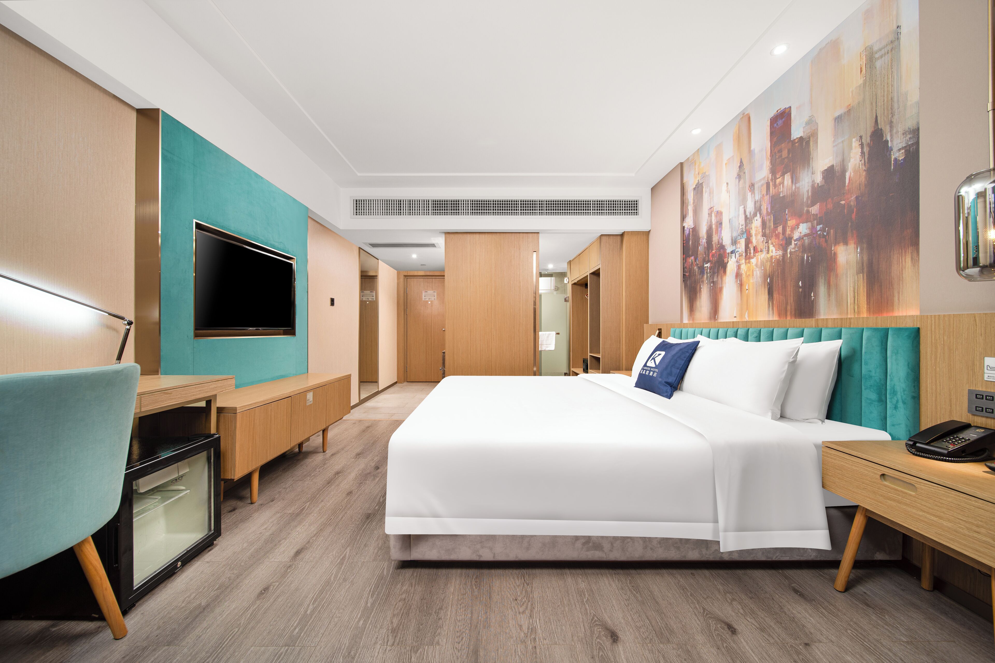 Une chambre d’hôtel moderne avec un grand lit, un bureau équipé d’un ordinateur, une télévision et un tableau représentant un paysage urbain accroché au mur.