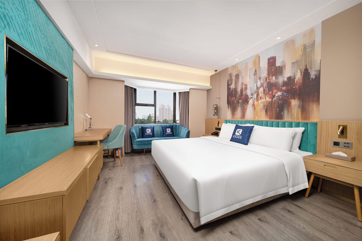A hotel room with a large bed, a desk, a chair, a TV, and a cityscape painting on the wall.