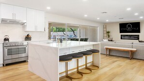 A modern kitchen with a central island, bar stools, a built-in oven, and a dining area with a view. - SWSS 8BR Palms Away Estate | Pool, Hot Tub, Sport Court, Putting Green & Games (Scottsdale)