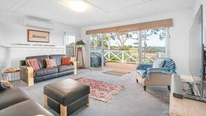 A spacious living area with a black leather sofa, a wooden coffee table, and a beige armchair. There is a large window with a view of the outdoors, a framed picture on the wall, and a small table with a plant. - Mandalay on Pipeclay - 3 Bedroom House - Free Parking - Free WIFI (Cremorne)