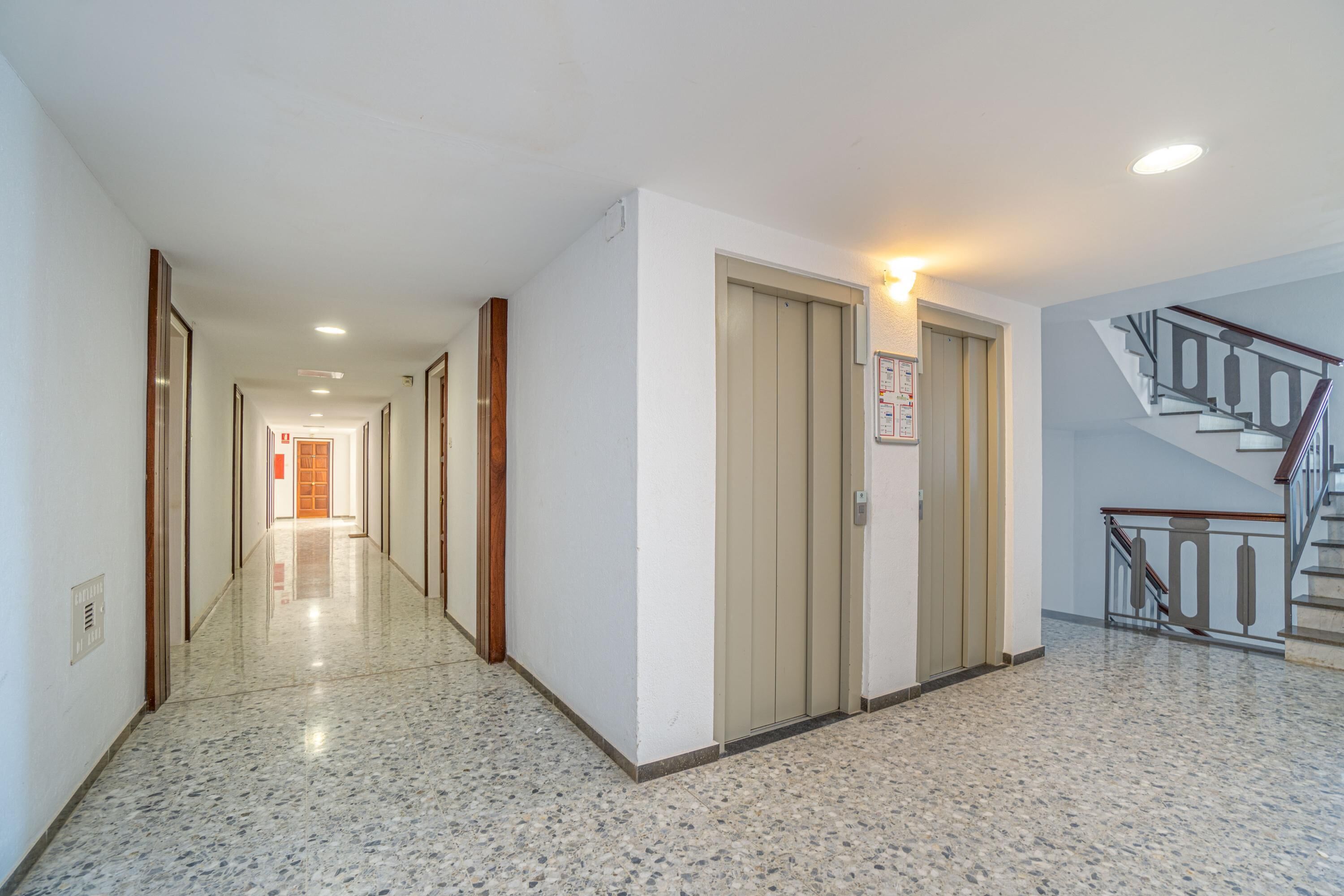 A long, well-lit hallway with a polished floor, wooden doors, and a staircase with a metal railing.