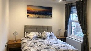 A bedroom with a bed, two bedside tables, a ceiling fan, a window with curtains, and a sunset painting on the wall. - Two Bedroom House, Birchington, ct79rn (Birchington)