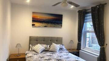 A bedroom with a bed, two bedside tables, a ceiling fan, a window with curtains, and a sunset painting on the wall.