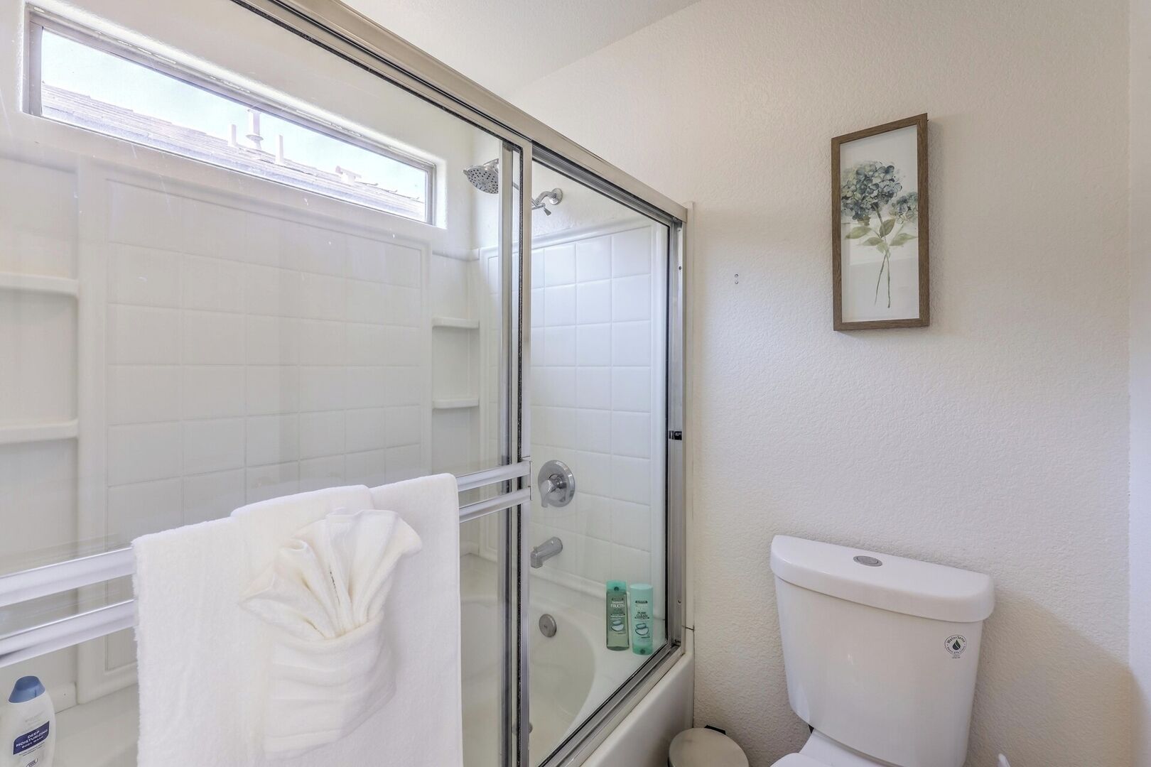 A bathroom with a glass shower enclosure, a white toilet, and a framed floral artwork on the wall.