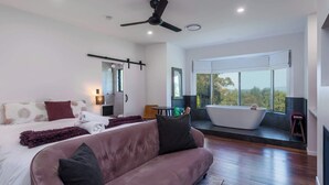 A modern bedroom with a large bed, a sitting area with a sofa, a bathtub, and a view of the outdoors.