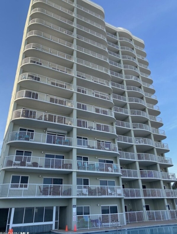 A multi-story hotel with balconies on each floor, a pool area at the bottom, and a clear sky.