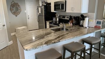 A kitchen with white cabinets, a granite countertop, a stainless steel refrigerator, a microwave, an oven, and a coffee maker. There are three bar stools and a welcome sign on the wall.