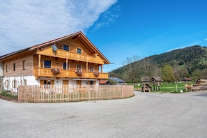 A wooden chalet with a balcony, a fenced driveway, and a mountainous backdrop. - Apartment "Brauneck" with Mountain View, Balcony and Wi-Fi (Gaißach)