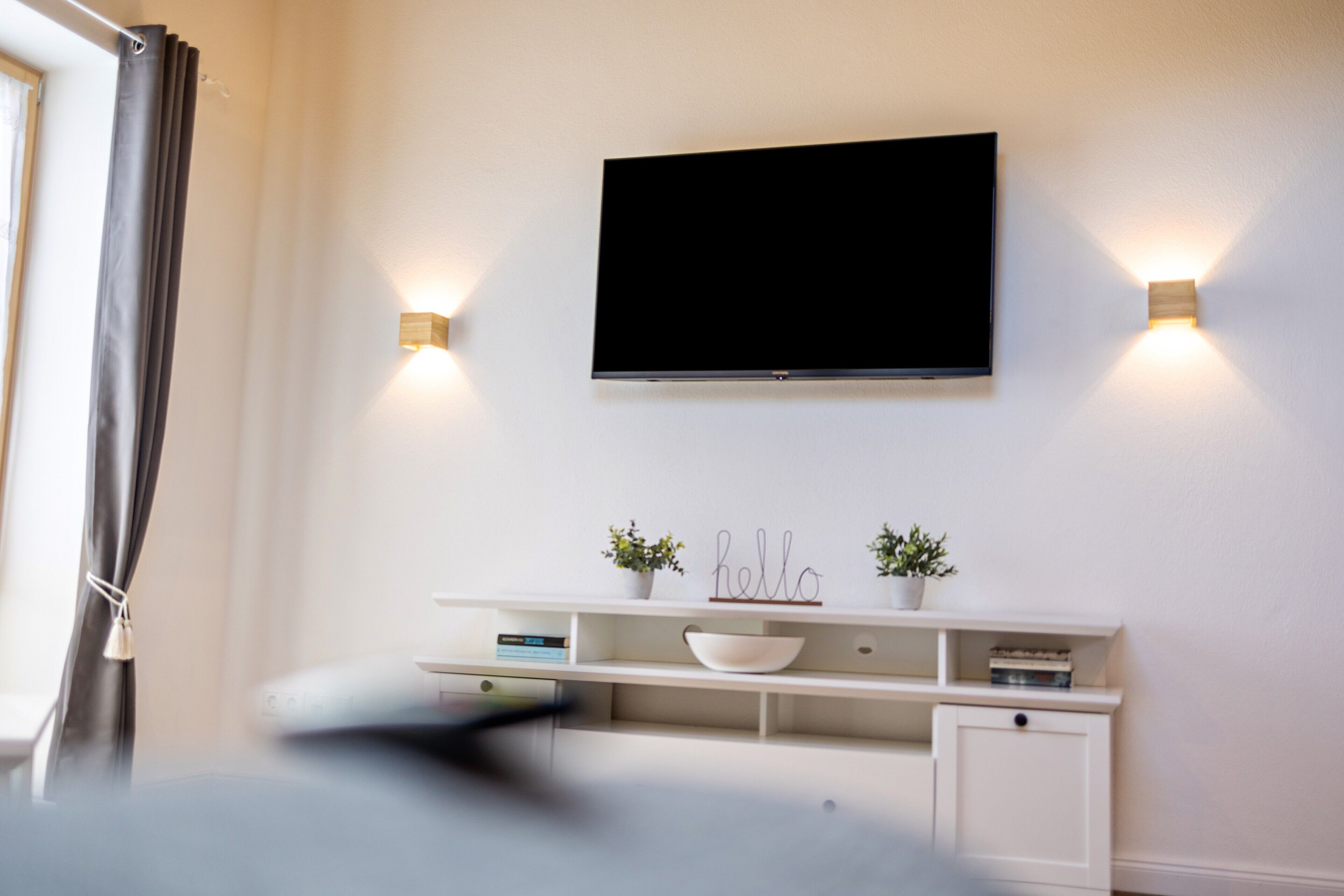 Ein modernes Zimmer mit einem Wand-Fernseher, einem weißen Konsolentisch mit Dekoration und zwei Wandleuchten.