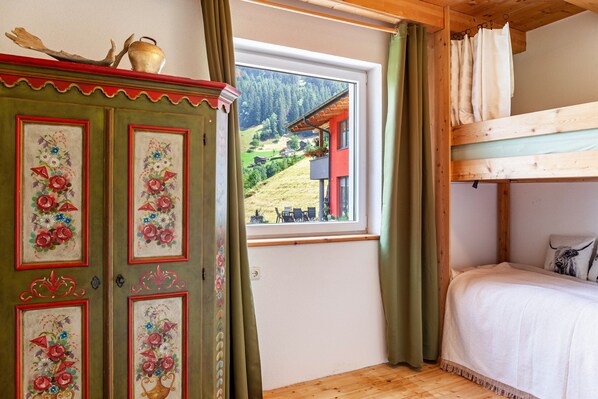 Una habitación con una cama marinera de madera, un armario verde con motivos florales y una ventana que da a una casa y montañas.