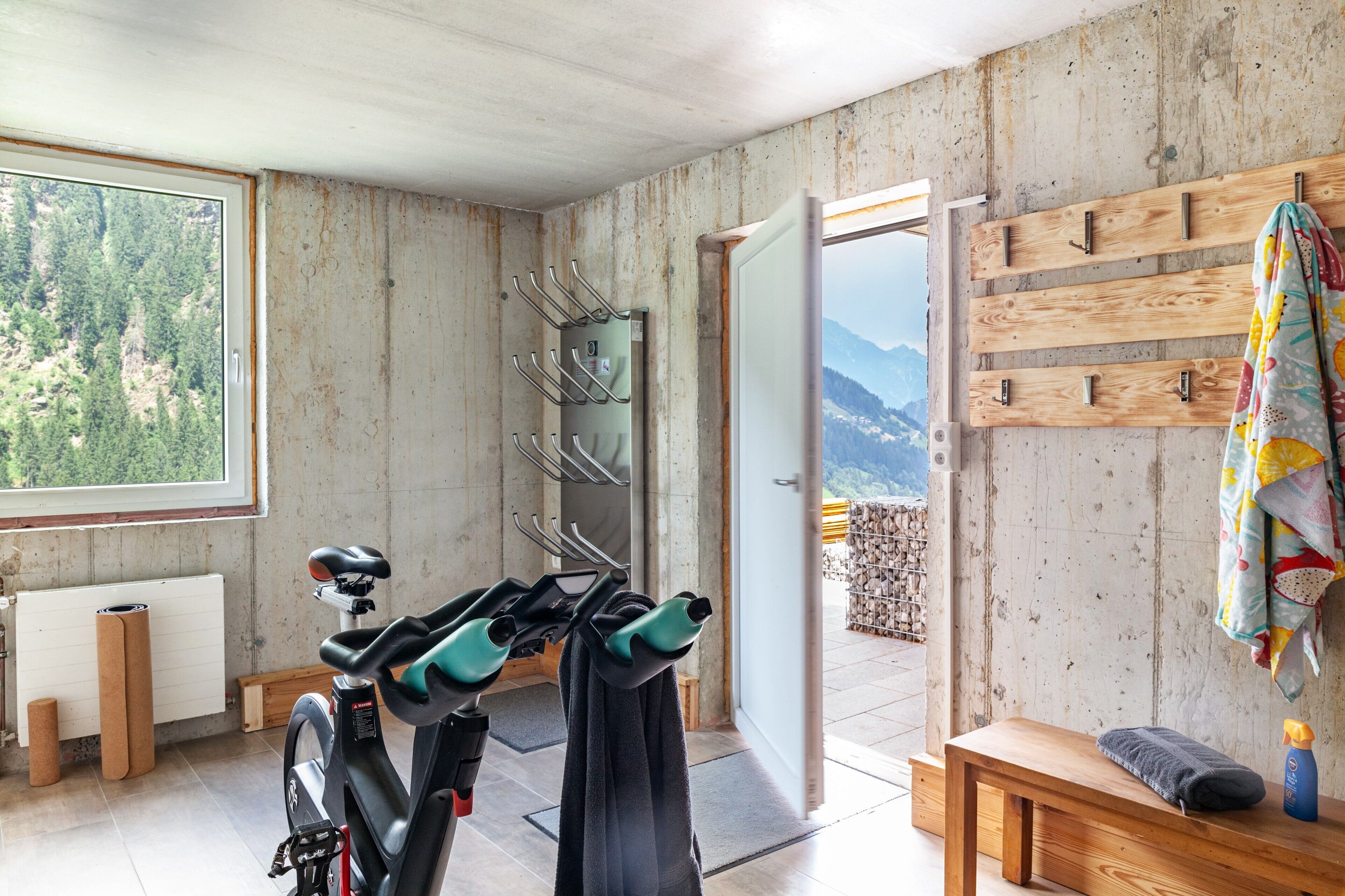Quarto com bicicleta, um banco, uma estante de madeira fixada na parede, e vista para montanhas através de uma janela grande.