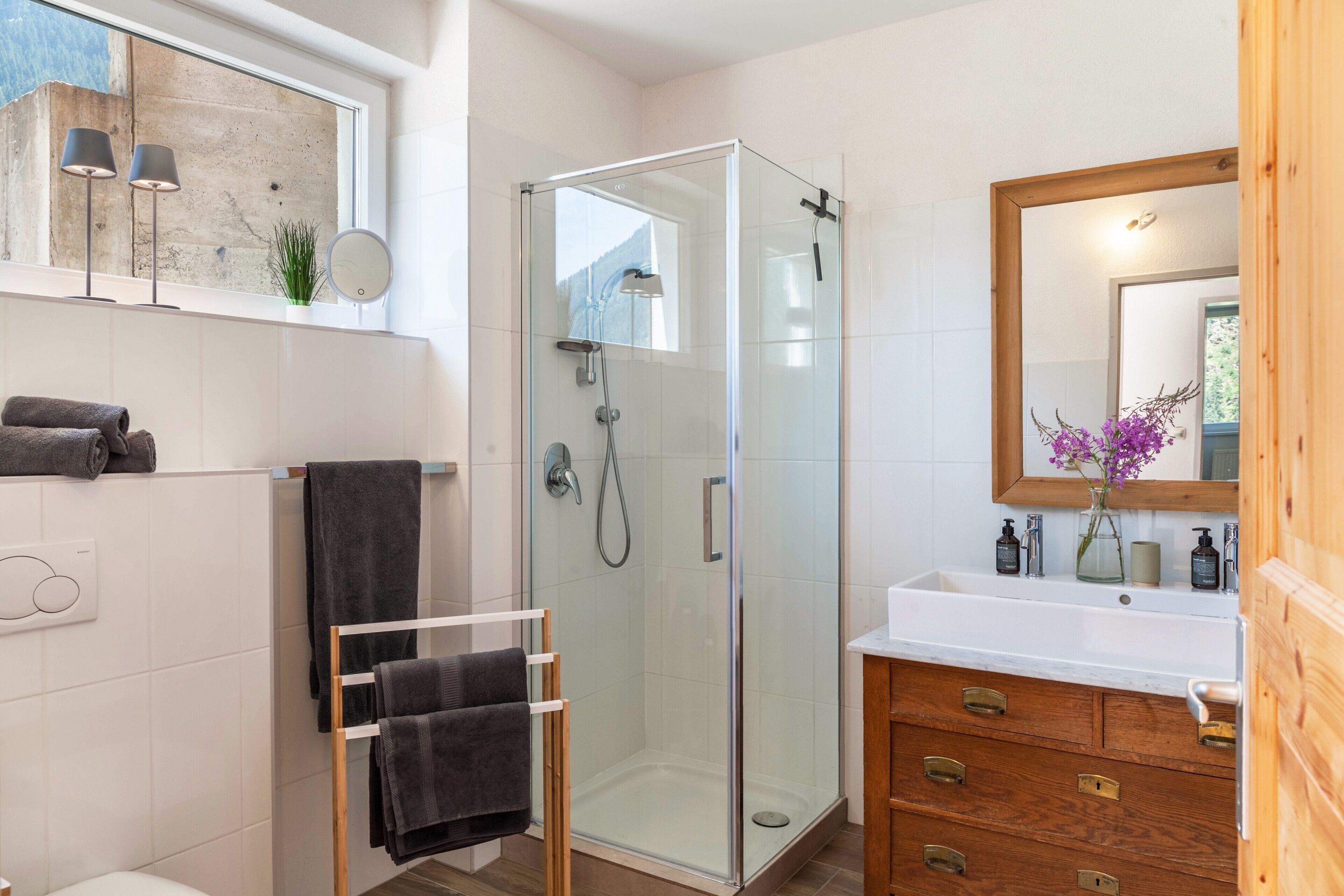 A modern bathroom with a glass shower enclosure, a white sink with wooden cabinetry, and a mirror with a vase of flowers.