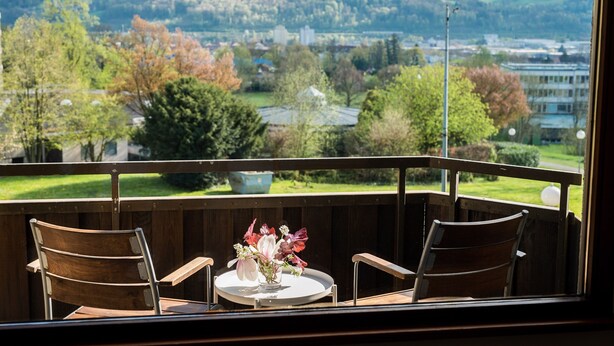 A balcony with two wooden chairs, a small round table, and a vase of flowers. A view of a town with buildings and trees is visible through the window.