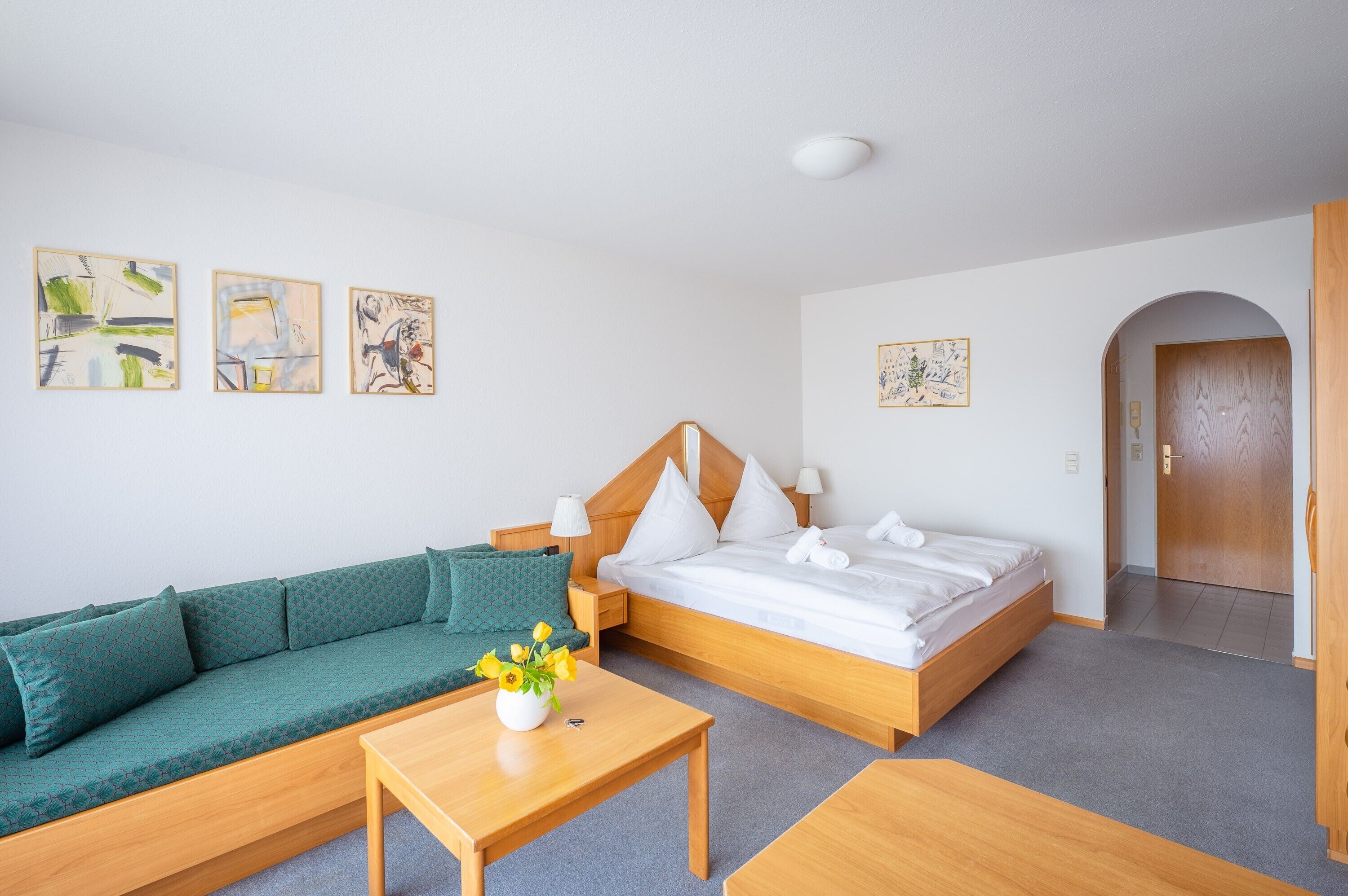 Hotelkamer met een bed, een groene bank, een houten tafel met een bloevas en kunstwerken aan de muur.
