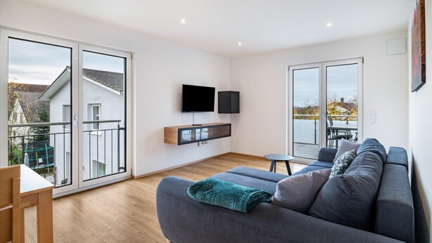Een moderne woonkamer met een grijze bank, een flatscreen televisie aan de muur en een balkon met uitzicht.