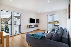 Un salon moderne avec un canapé gris, un téléviseur à écran plat fixé au mur et un balcon offrant une vue sur l’extérieur.