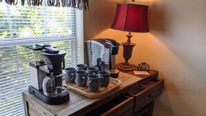 Una estación de café con cafetera, tazas y una lámpara sobre una mesa de madera cerca de una ventana con persianas.