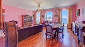Une pièce avec un plancher en bois, une table à manger entourée de chaises, un canapé, un lustre et de grandes fenêtres avec des rideaux.