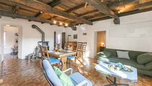 Un interior rústico con vigas de madera a la vista, una zona de comedor con mesa y sillas, un sofá verde y una mesa blanca con una bandeja de objetos.