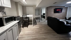 Eine moderne Küche mit weißen Schränken und einer eingebauten Mikrowelle, ein Essbereich mit weißem Tisch und Stühlen sowie eine Wohnarea mit einer schwarzen Couch und einem Fernseher, der 'NETFLIX' anzeigt.