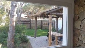 A view through a window of a patio area with a wooden table and chairs, a thatched roof structure, and a stone wall.
