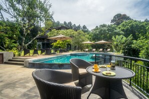 Zona de piscina con mobiliario de mimbre y sombrillas, rodeada de exuberante vegetación y un edificio al fondo.