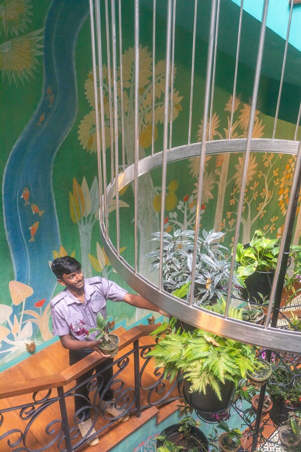 A person standing on a wooden staircase with a railing, surrounded by potted plants and a mural with floral and tree designs. - Kandy Dendrobium House (Puliyadda)