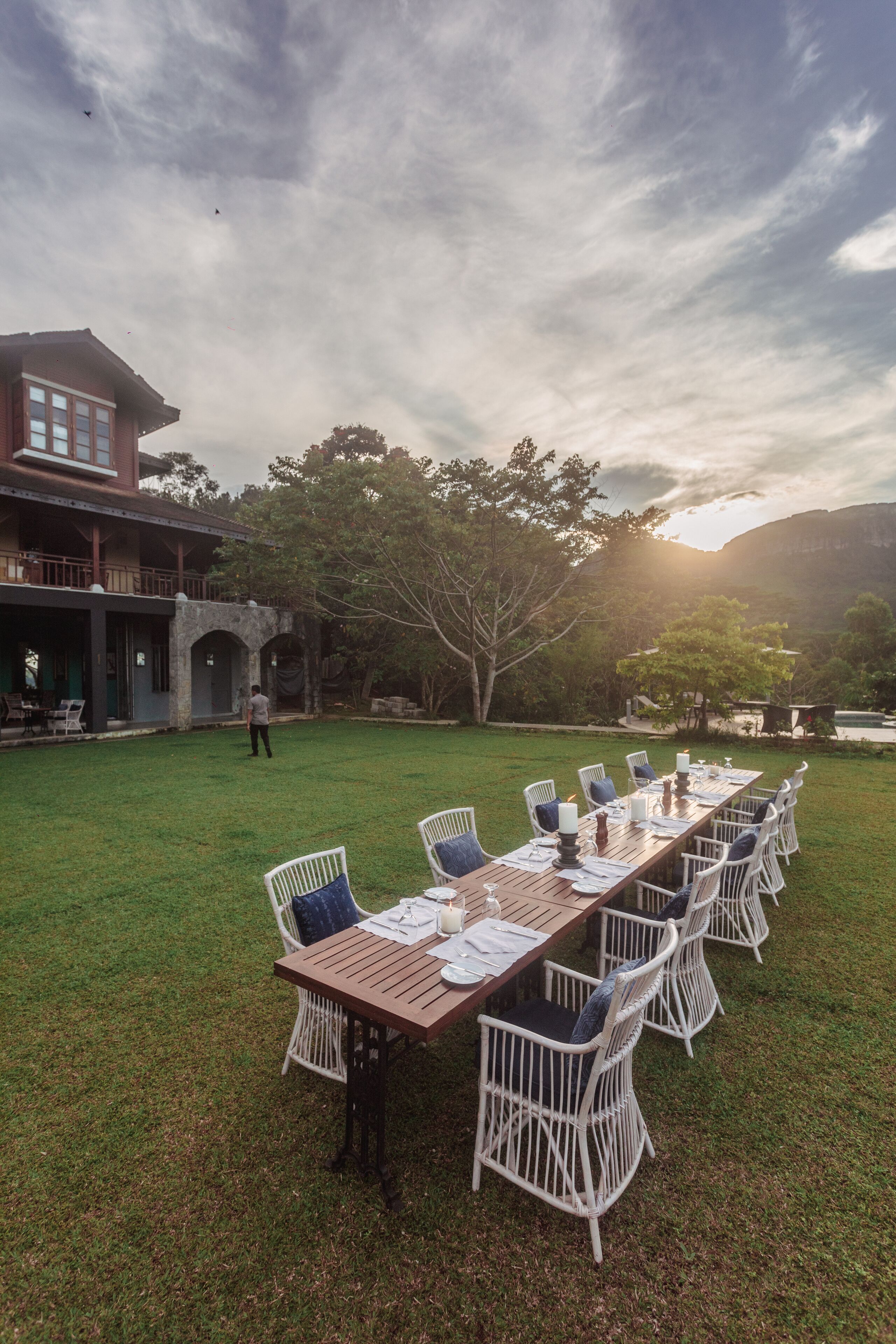 An outdoor dining area with a long wooden table and white wicker chairs, set against a backdrop of a building and trees under a cloudy sky.