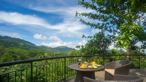 A balcony with wicker furniture, a table set with yellow cups and a glass, and a scenic view of green hills and a clear sky. - Kandy Dendrobium House (Puliyadda)