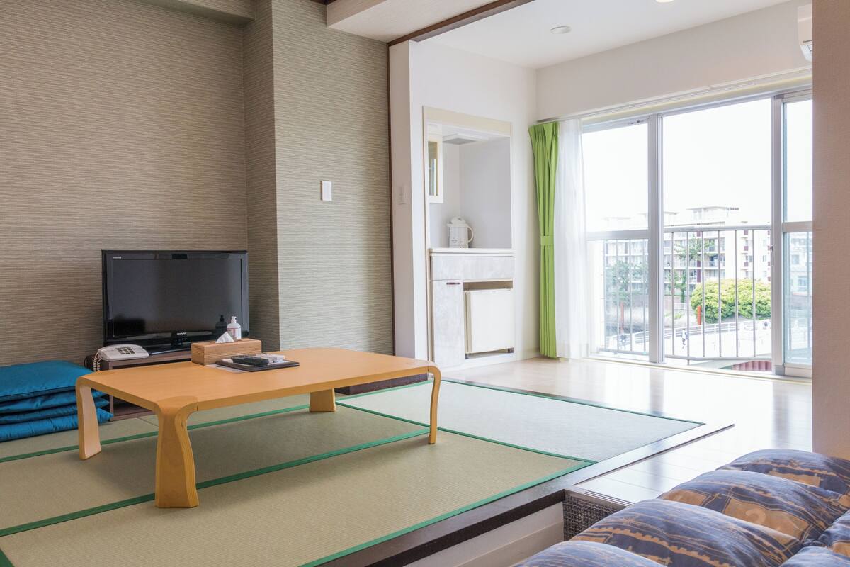 A room with tatami flooring, a television, a table, a sofa, and a view through a large window.