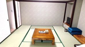 A tatami mat room with a wooden table, a small shelf with items, and a TV on a stand. - kishu railway katase enoshima hotel (Fujisawa)