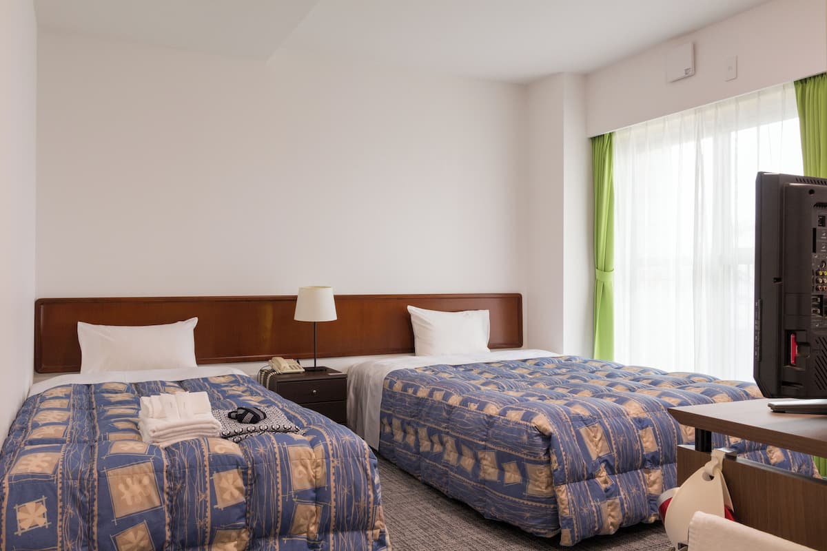 A hotel room with two beds, a desk, a TV, and a window with curtains.