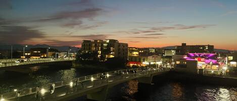 Une vue urbaine le long du littoral, avec des bâtiments illuminés, un pont et une rivière.