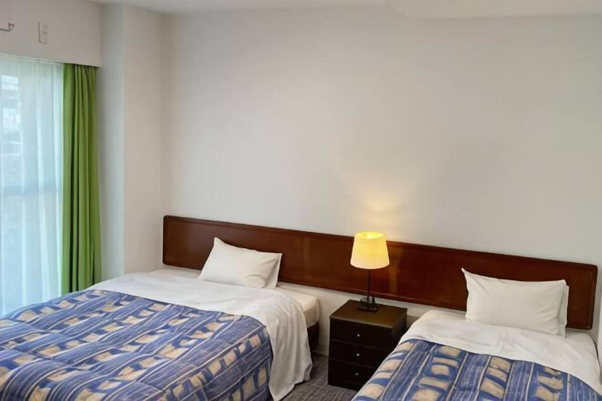 A hotel room with two beds, a wooden headboard, a nightstand with a lamp, and a window with green curtains.
