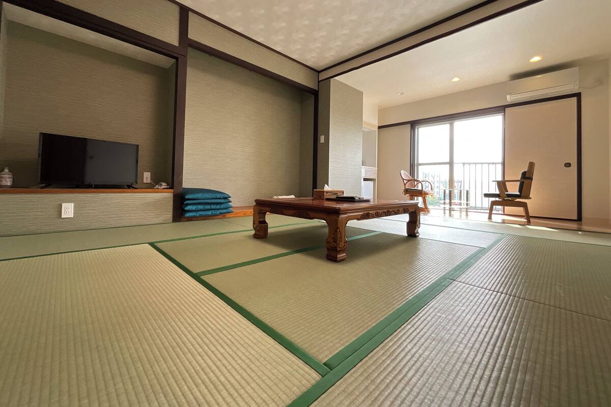 A traditional tatami mat room with a wooden table, a television on a shelf, and a sliding door leading to a balcony.