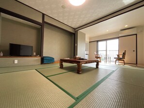 Una habitación tradicional con esteras de tatami, una mesa de madera, un televisor sobre una estantería y un puertas correderas que dan a un balcón.