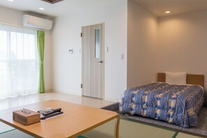 Una habitación de hotel con una cama, una mesa con una caja de pañuelos y controles remotos, una cortina verde y una puerta.