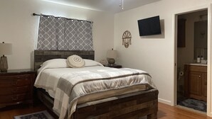 A bedroom with a wooden bed, a ceiling fan, a TV, and a bathroom visible through an open door.