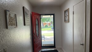 A hallway with a red door, a black mat at the entrance, and a small window with a view of the outside.