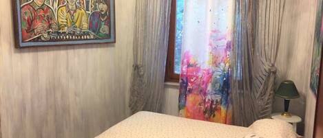 Un dormitorio con cama, un cuadro, una ventana con cortinas y una lámpara.