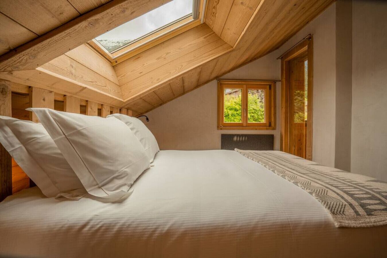 A bedroom with a skylight, a bed with pillows, a window, and a view of greenery outside.