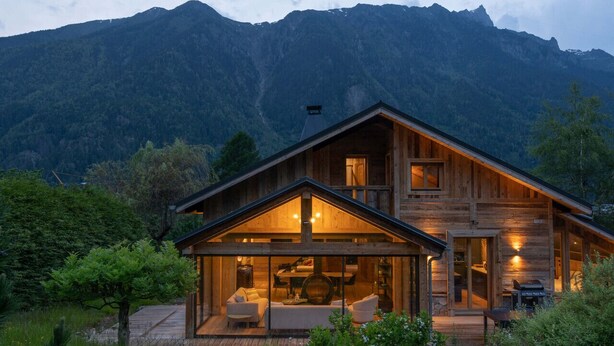 A wooden cabin with a large glass front, a deck, and a mountain backdrop.
