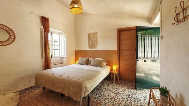 Une chambre avec un lit, une table de chevet avec une lampe, une décoration murale en macramé, une armoire en bois et une fenêtre avec des rideaux.