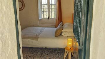 Een slaapkamer met een bed, een raam met gordijnen, een hanglamp en een klein tafeltje met een lamp.