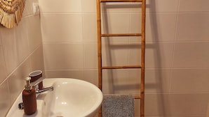 Une salle de bain avec une échelle en bois appuyée contre le mur, un lavabo blanc avec un distributeur de savon et un miroir doté d’un cadre tressé.