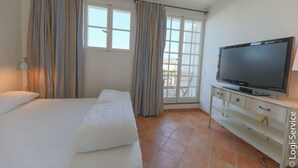 A hotel room with a bed, a TV on a stand, a window with curtains, and a view of the outside. - Air-conditioned house, close to beach 9 people  et  mooring (PORT GRIMAUD)