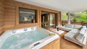 Eine Holzterrasse mit Whirlpool, einem weißen Sofa und einer Glastür, die zu einem Außenbereich führt.