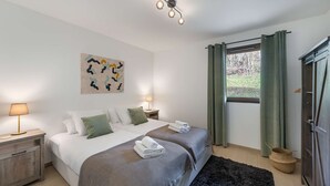 Ein Schlafzimmer mit zwei Betten, einem Nachttisch mit Lampe, einem Bild an der Wand, einem Fenster mit Vorhängen und einem Holzkasten.