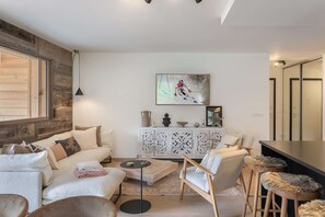 A modern living room with a sofa, coffee table, and bar stools. There is a cabinet with decorative items and a painting on the wall.
